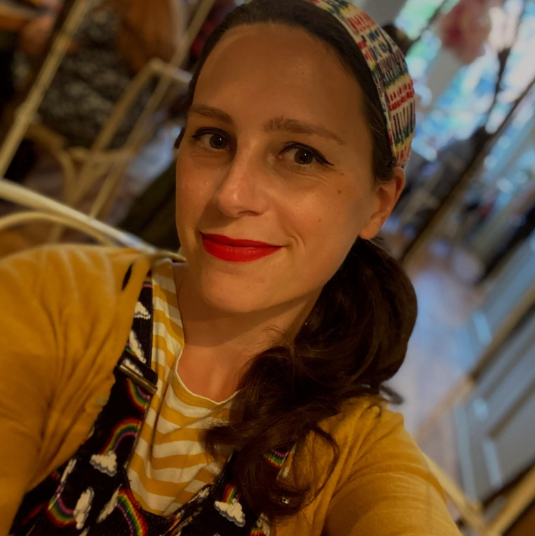 Rosa Lee wearing a mustard yellow cardigan over a yellow and white striped shirt, a black patterned dress, and a colorful patterned headband, captured in a warm, slightly blurry indoor setting.